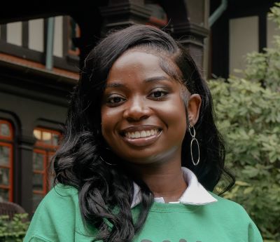 A woman wearing a green sweater is smiling. In the background is a building with windows.